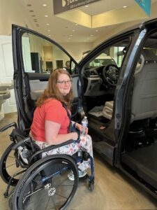 A woman in a wheelchair, smiling and holding a water bottle, sits next to an open car door in a showroom. The car appears to have accessibility modifications for wheelchair users.