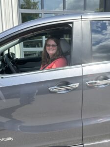 A woman with glasses and long hair, wearing a red top, smiles while sitting in the driver’s seat of a gray car parked outside on a sunny day.