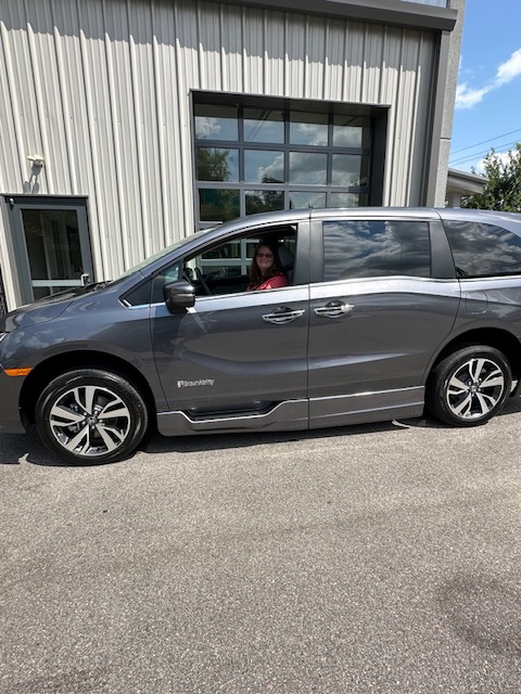 A person sits in the driver’s seat of a gray minivan parked outside a building with a large garage door and metal siding. The weather is sunny with a partly cloudy sky reflected in the windows.