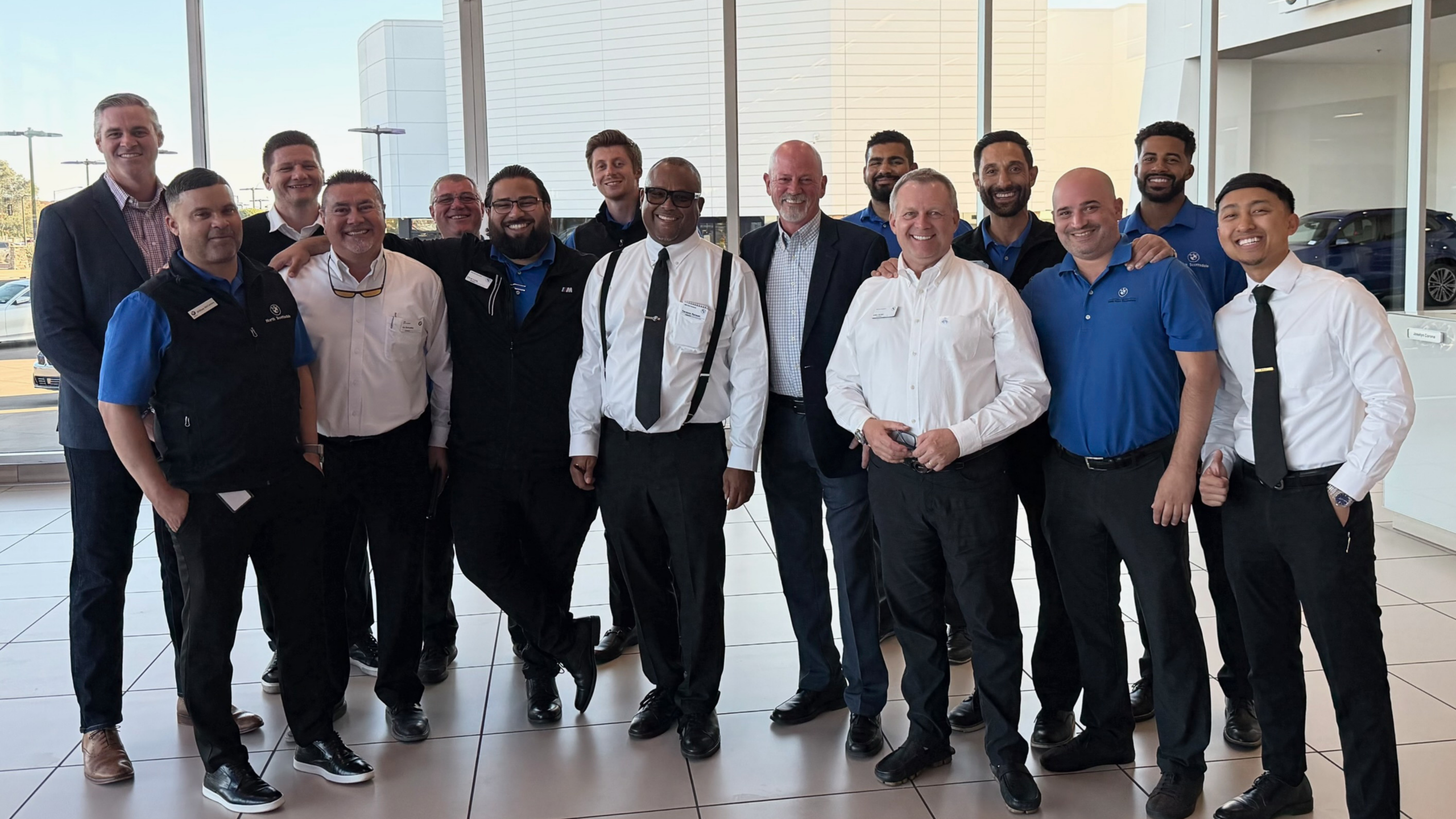 A group of fifteen men, wearing business and business-casual attire, stand together inside a modern, bright showroom, smiling at the camera. Large windows and cars can be seen in the background.