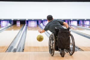A person in a wheelchair bowls with a yellow ball in a bowling alley, aiming toward a set of pins. The image is taken from behind, capturing the action and the lanes ahead.
