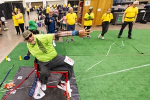 An older man in a neon green shirt throws a discus from a seated position at an indoor sporting event. Spectators and staff in yellow shirts watch from behind on artificial turf. The scene is energetic and focused.