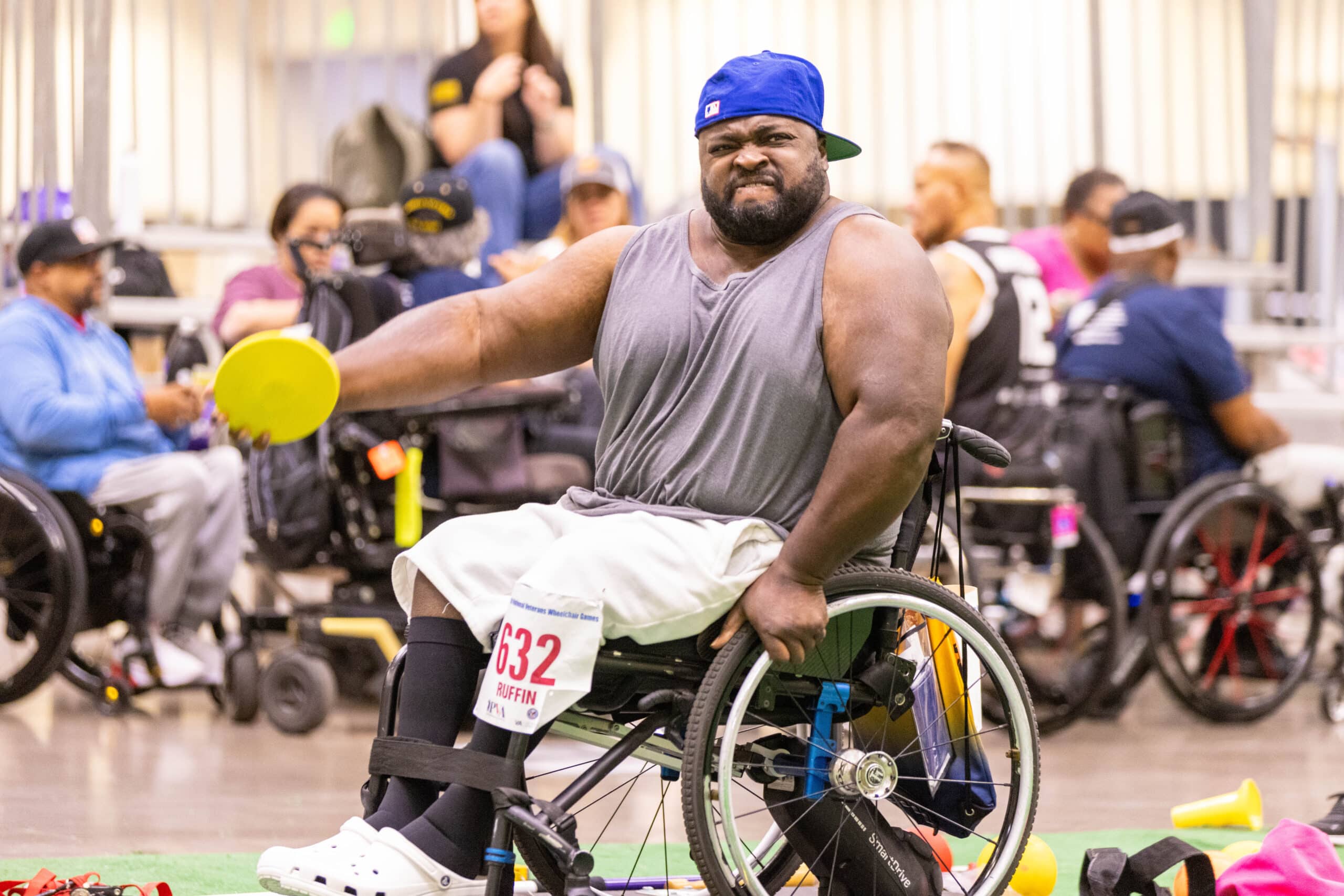 A man in a wheelchair wearing a blue cap, gray tank top, and white pants holds a yellow disc in a gym setting. He participates in a sporting event with other wheelchair users in the background.