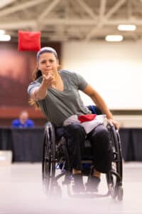 A woman in a wheelchair throws a bean bag during an indoor game. She is focused, wearing a gray shirt and black pants, with another bean bag resting on her lap. The background is blurred.