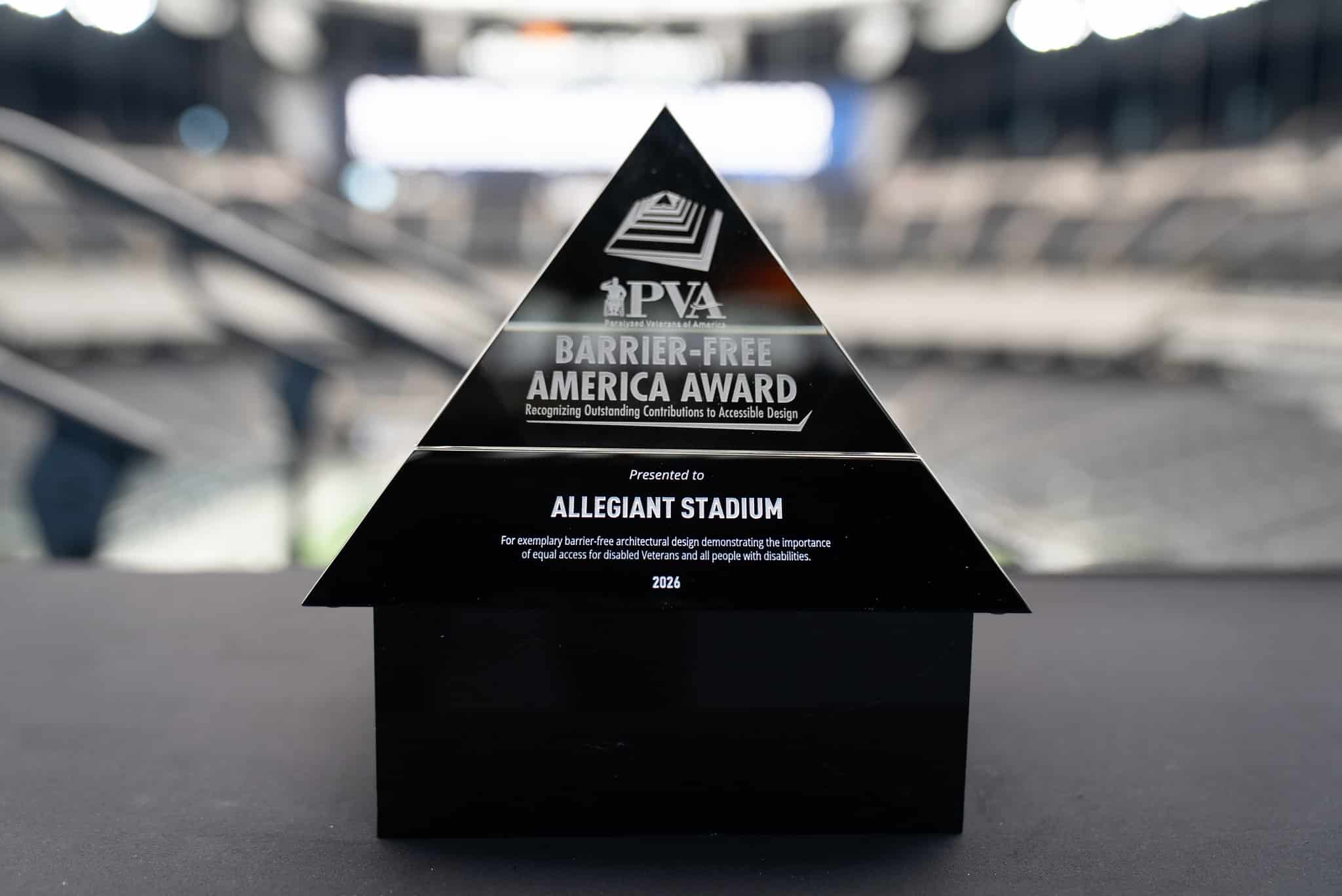 A triangular black trophy with the inscription "PVA Barrier-Free America Award" recognizing Allegiant Stadium for outstanding contributions to accessible design, displayed indoors with a blurred background.