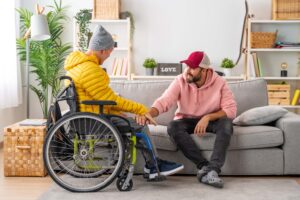 Two men smiling and holding hands; one is sitting on a sofa and the other is in a wheelchair. They are indoors in a cozy, bright living room with plants and books on shelves in the background.