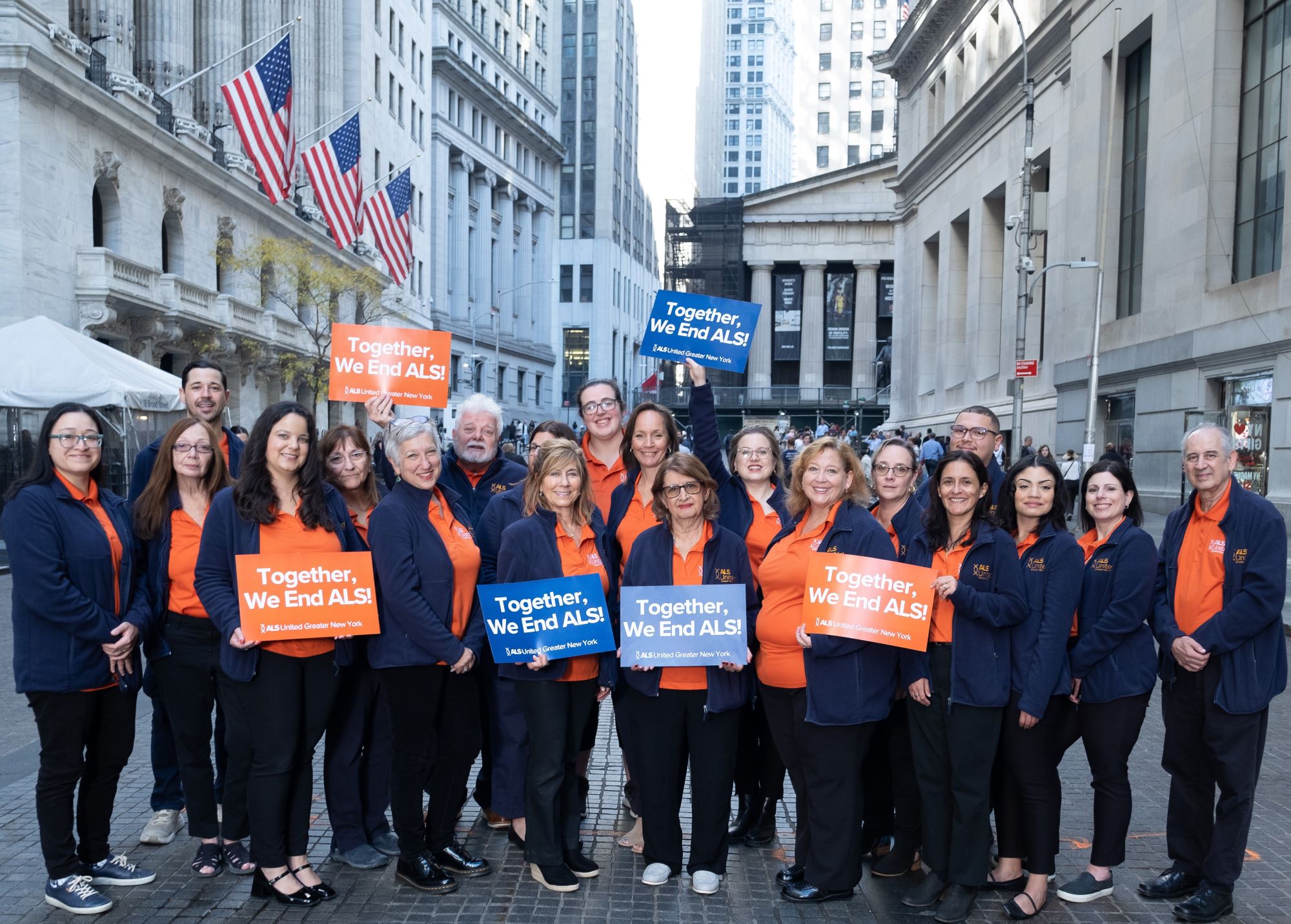 A large group of people stand together on a city street, wearing matching orange and navy shirts and jackets, holding signs that read "Together, We End ALS!" and smiling at the camera. American flags hang in the background.