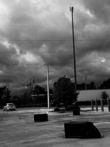 A black and white photo of an empty parking lot with two toppled trash bins, a single parked white car, cloudy sky, power lines, and a tall cell tower in the background.