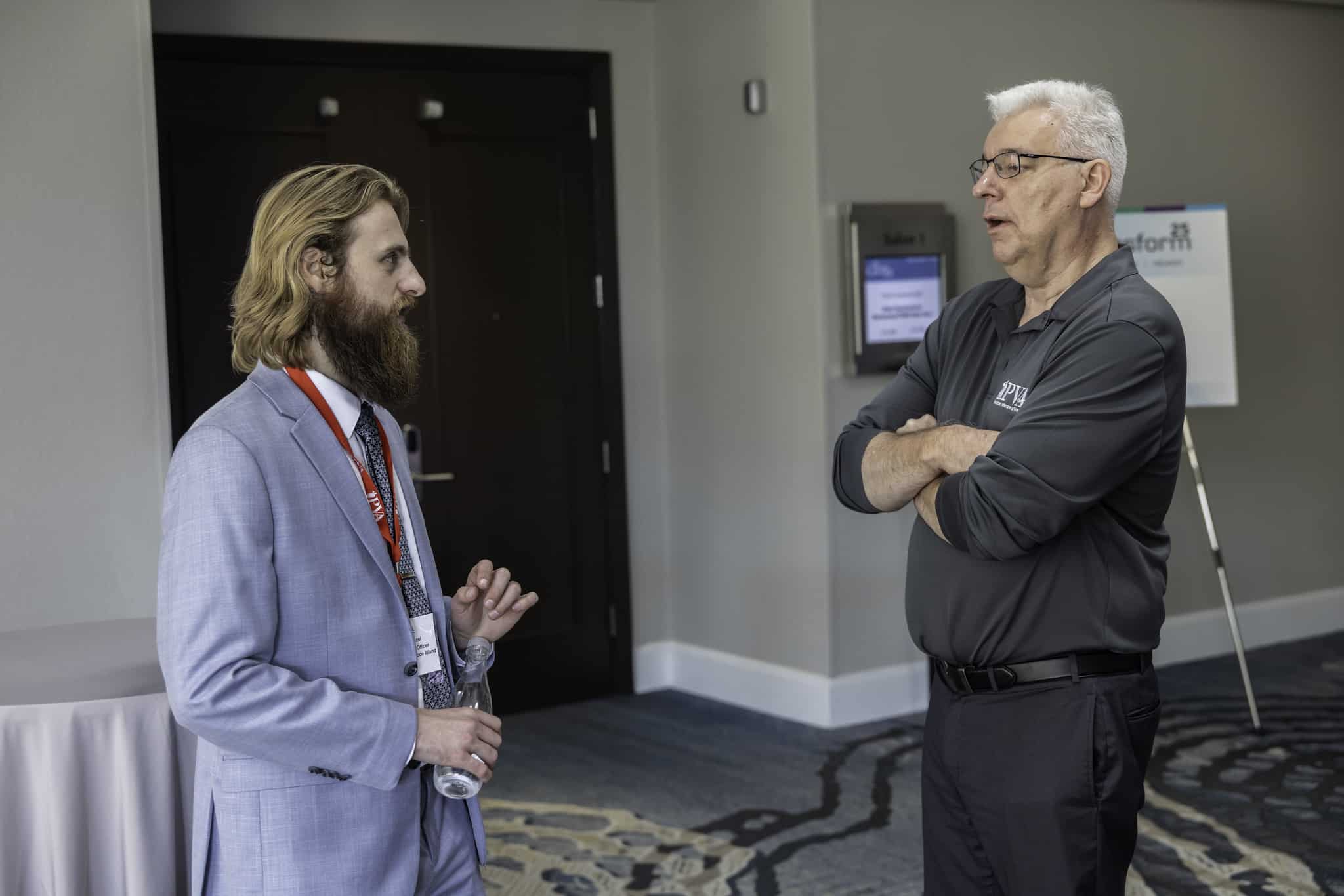 Two men stand indoors having a conversation. One man, in a light blue suit with a red lanyard, holds a water bottle, while the other, in a dark polo, stands with arms crossed. A signboard and closed doors are in the background.