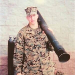 A person in military camouflage uniform and cap stands against a brick wall, carrying a large, tube-shaped object on their shoulder. Two tall metal cylinders are in the background.