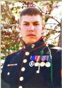 A young man in a U.S. Marine Corps dress uniform stands outdoors, wearing several medals and ribbons on his chest, with trees and sunlight in the background.