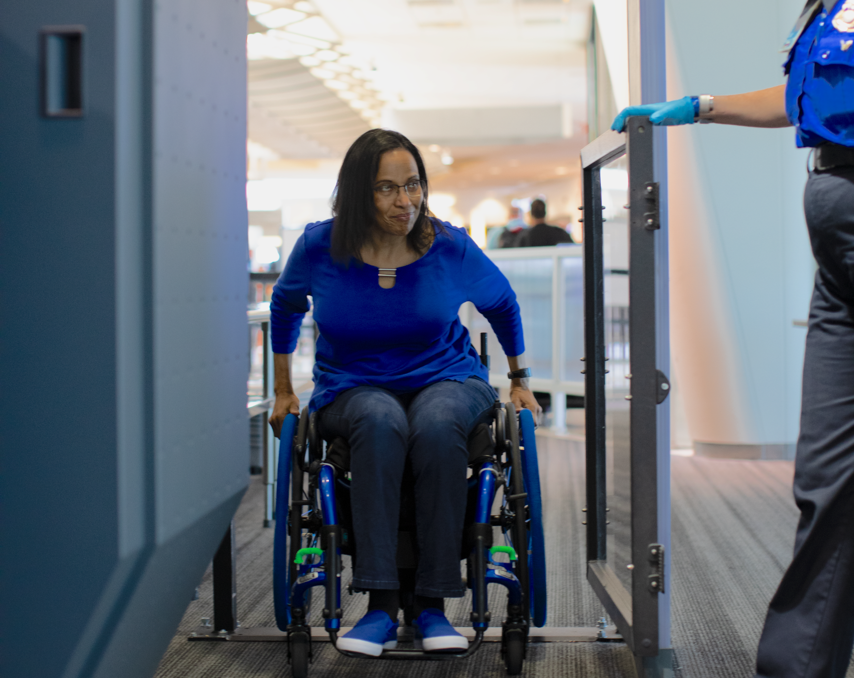 A woman in a wheelchair, dressed in a blue top and jeans, moves through an open security gate as a TSA officer in gloves stands nearby at an airport security checkpoint.