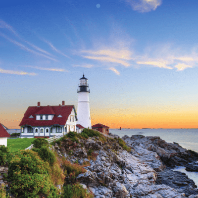 A white lighthouse with an attached house sits on a rocky shore at sunrise, with an American flag nearby, blue sky, wispy clouds, and calm ocean water in the background.
