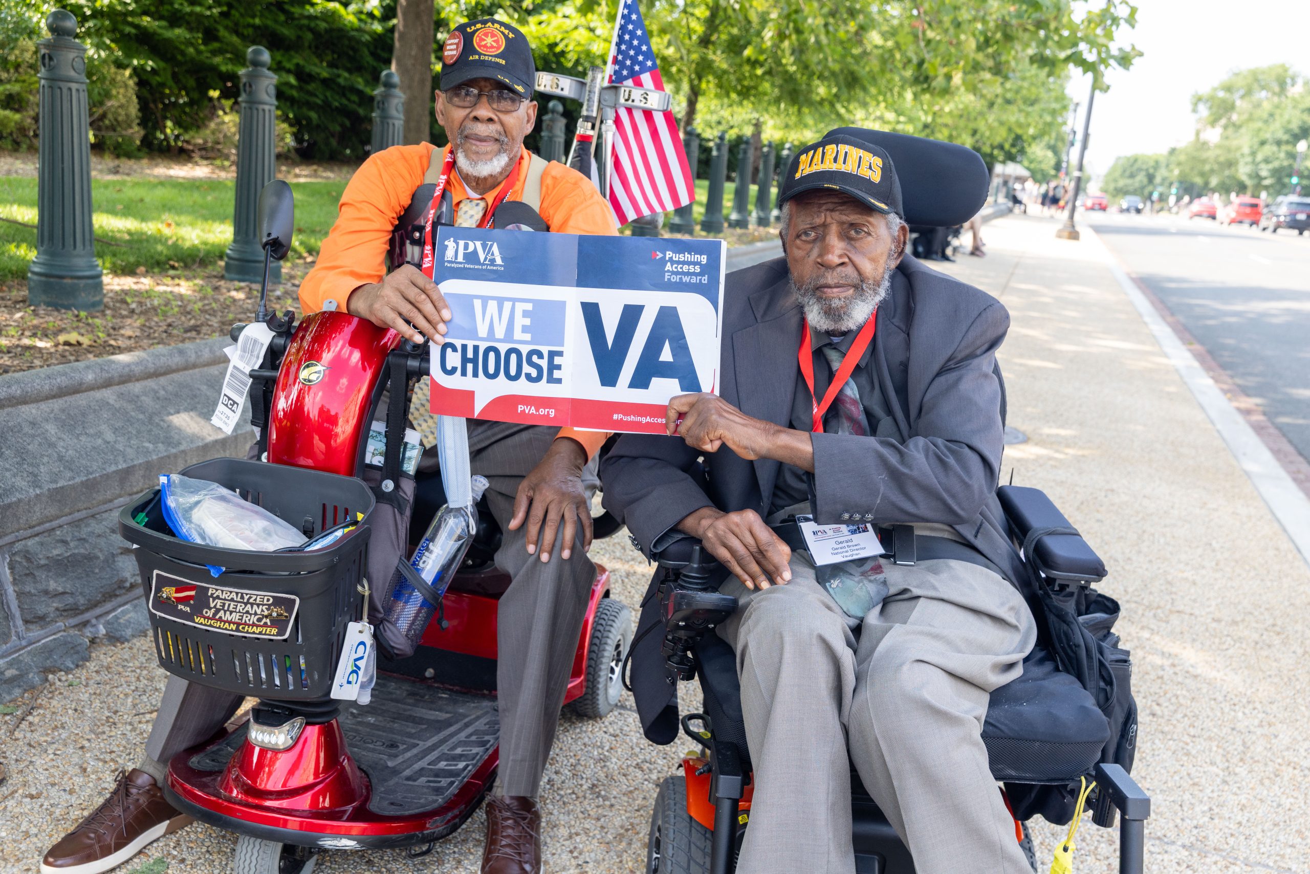 Two elderly men in wheelchairs hold a "We Choose VA" sign and wear military hats and badges. They are outdoors on a sidewalk, with trees and an American flag in the background.