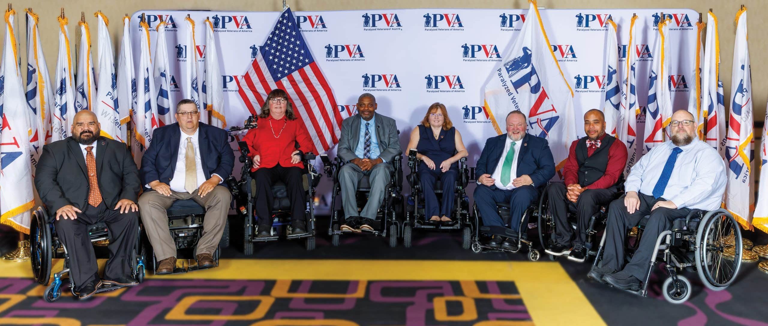 A group of eight people, some in wheelchairs, pose in front of a PVA-branded backdrop with American and organizational flags displayed behind them on a patterned carpet.