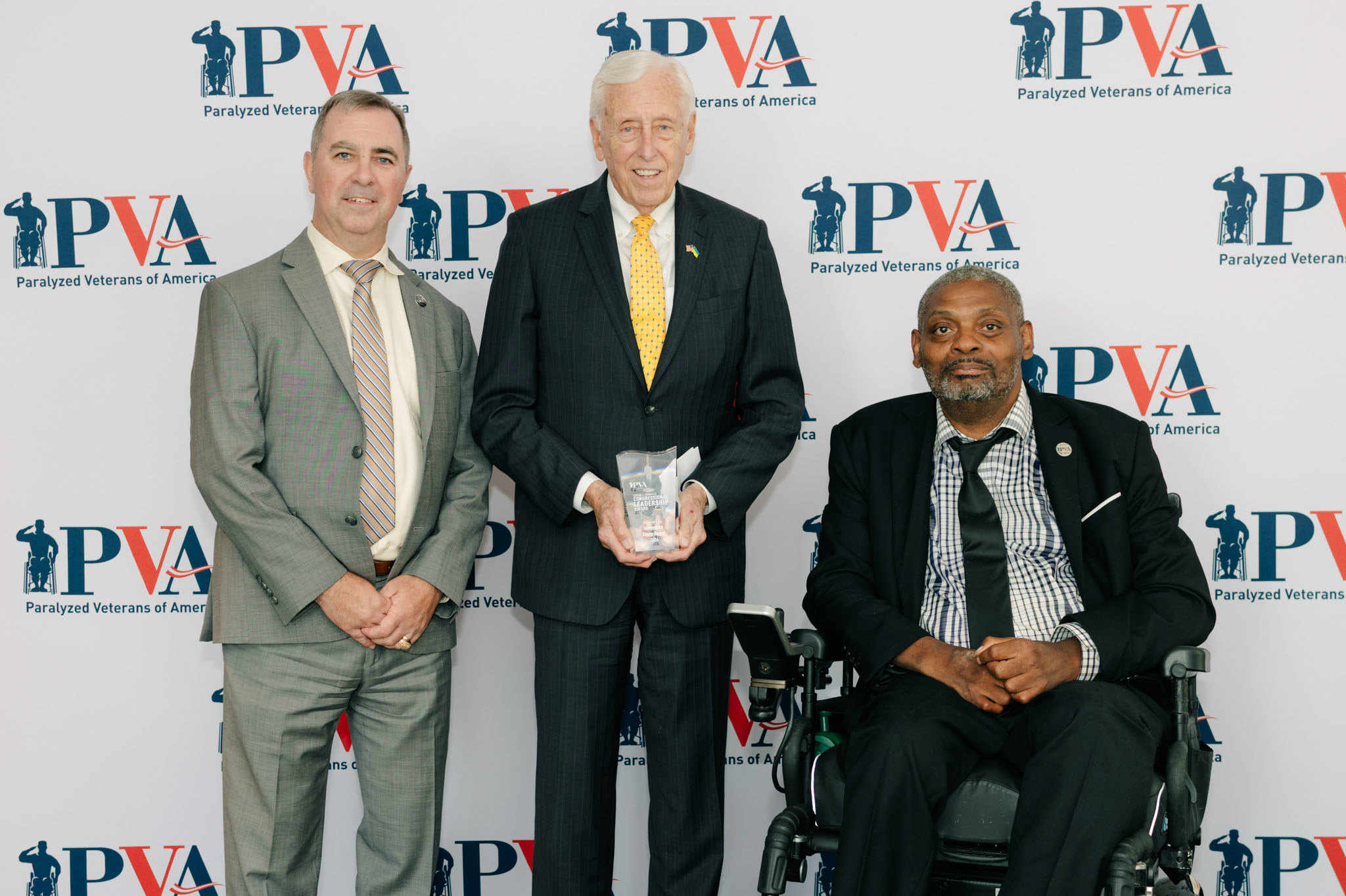 Three men pose for a photo at a Paralyzed Veterans of America (PVA) event; one man holds an award, another stands to his right, and a third man sits in a wheelchair on his left, all smiling in front of a PVA backdrop.