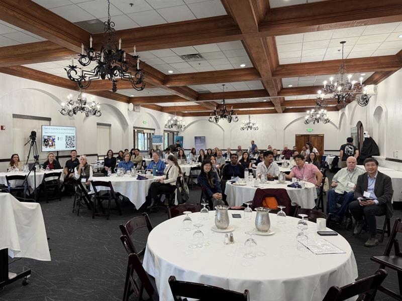 A group of people sit at round tables in a banquet hall, facing forward and listening attentively. The room has chandeliers, wooden beams, and white walls. Some people are taking notes or looking at laptops.