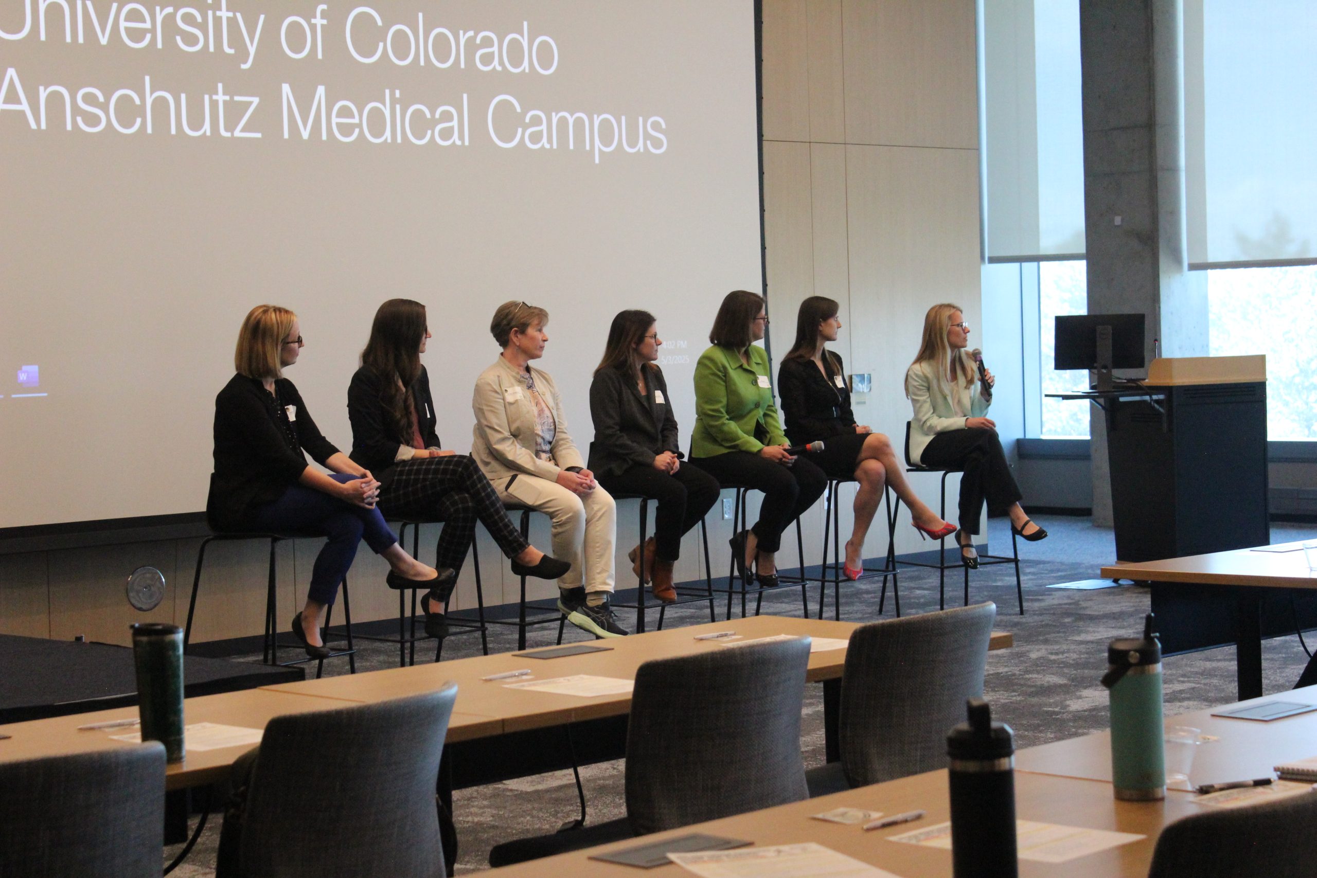 Seven women sit on stools in a row at the front of a conference room, with one speaking into a microphone. A projected screen behind them displays "University of Colorado Anschutz Medical Campus.