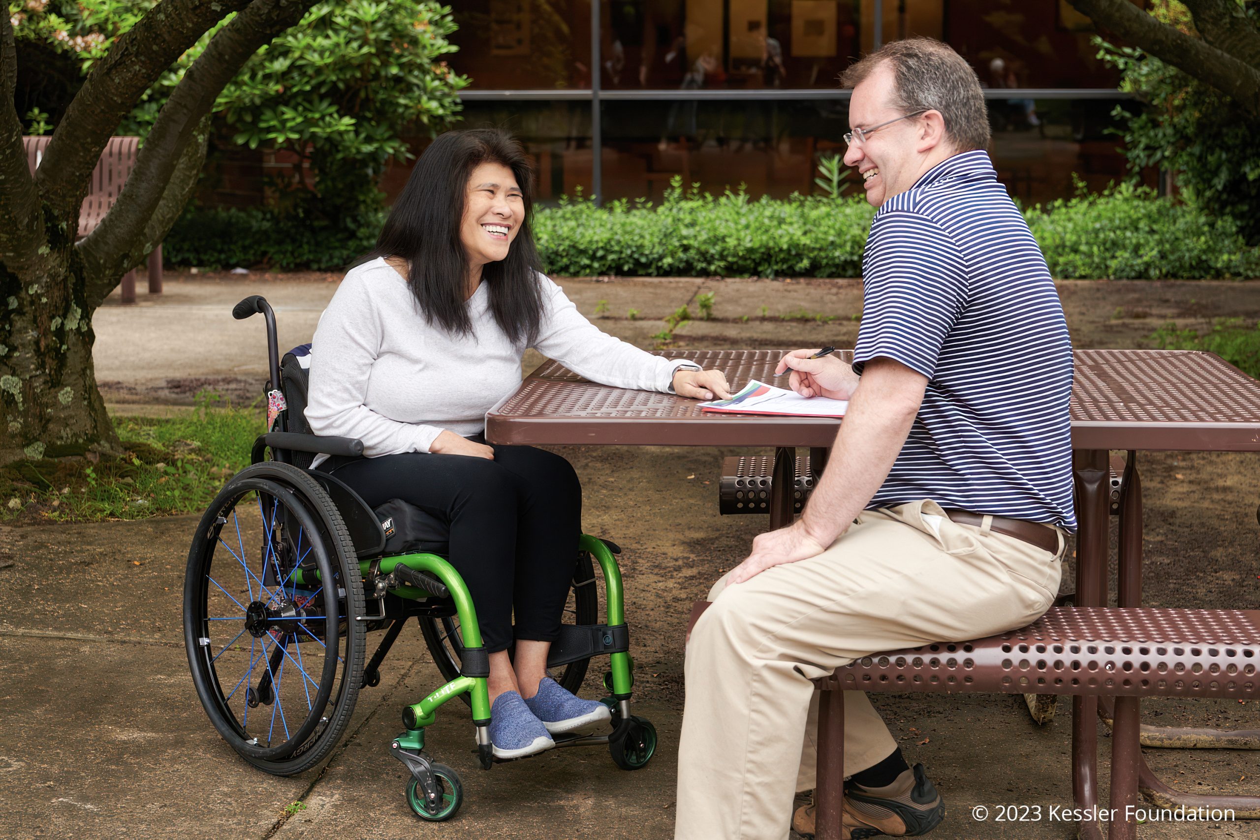 A woman in a wheelchair and a man sit outdoors at a picnic table, smiling and talking. Trees and bushes surround the area, and both appear relaxed and engaged in conversation.