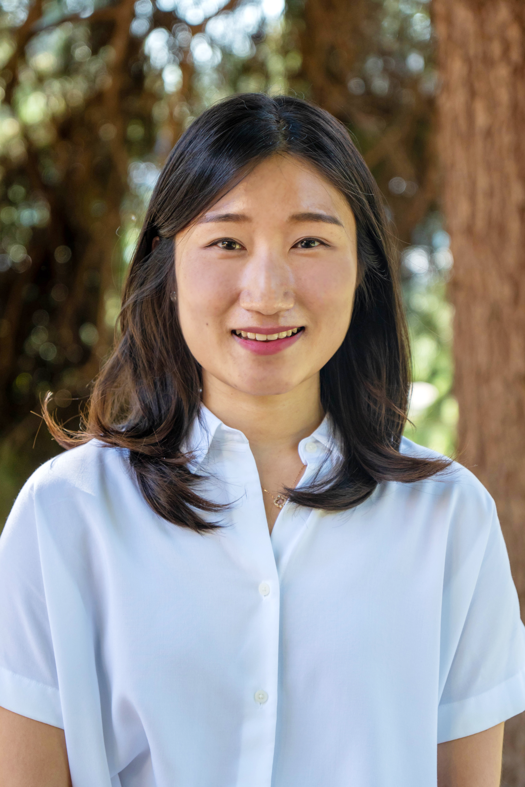 A woman with long dark hair, wearing a white button-up shirt, stands outdoors in front of blurred trees, smiling at the camera.