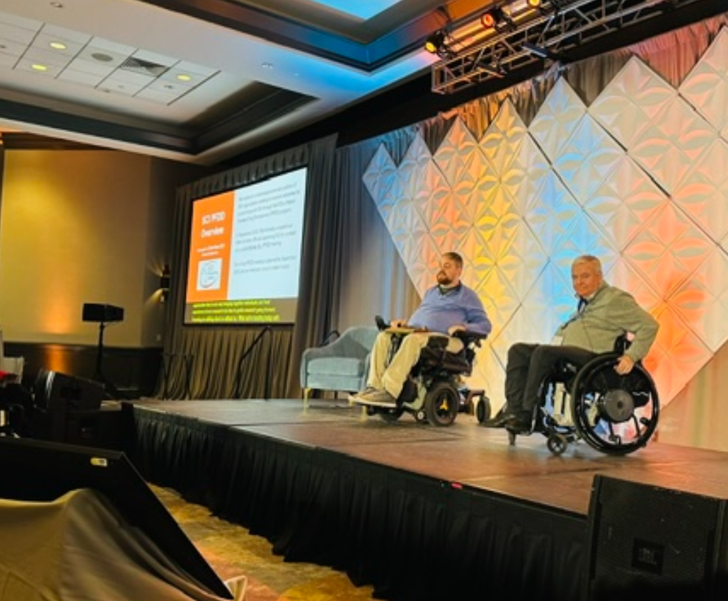 Two men in wheelchairs speak on stage at a conference. Behind them is a geometric backdrop and a large screen displaying a presentation slide. The room is well-lit with a professional setup.