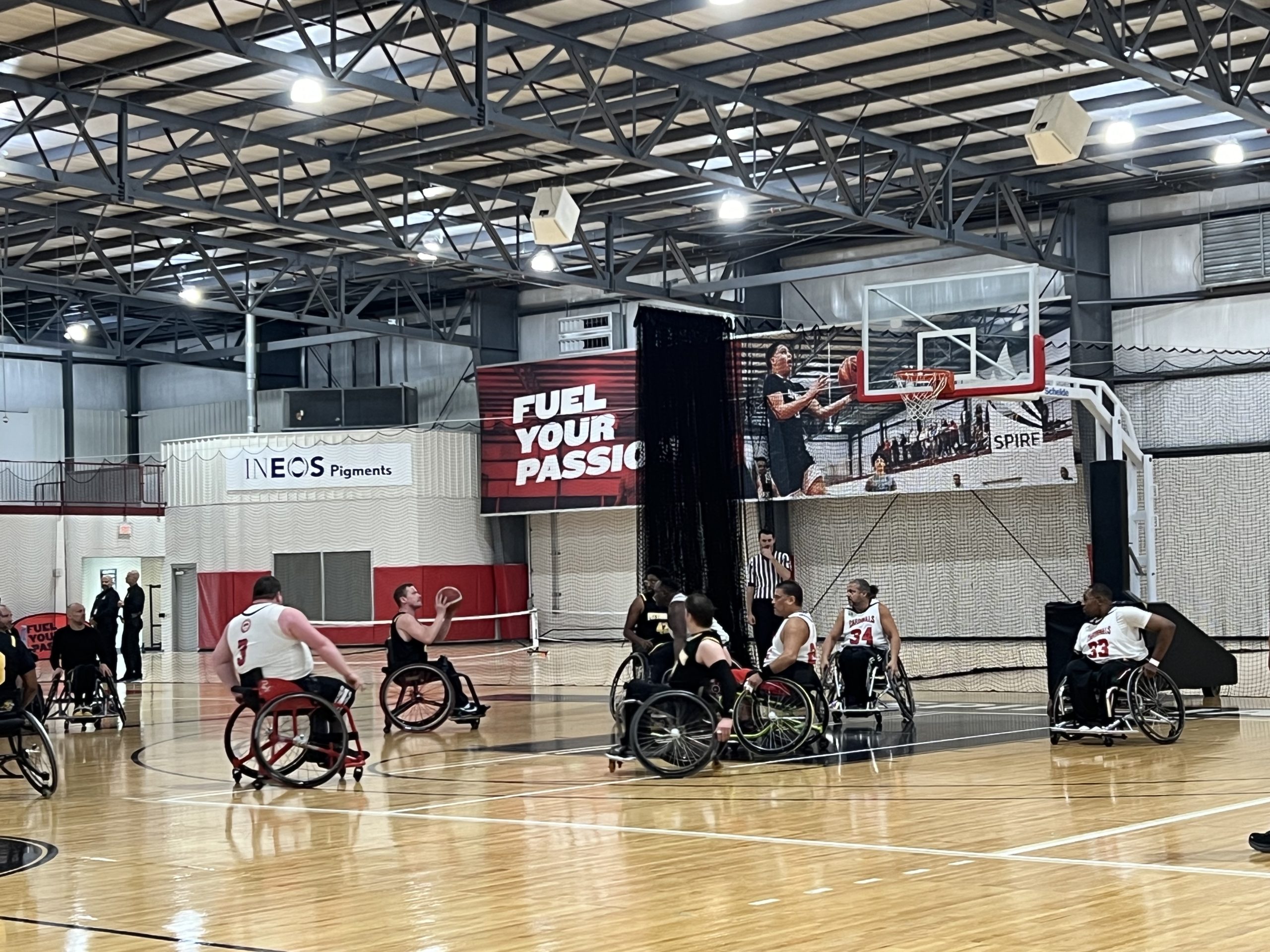Wheelchair basketball players compete on an indoor court, with some players preparing for a rebound near the basket. In the background, a large banner reads "FUEL YOUR PASSION." The gym is brightly lit.