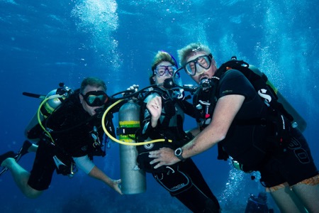 Three scuba divers underwater, wearing wetsuits and dive masks, gather around an oxygen tank and smile at the camera, surrounded by blue water and rising bubbles.
