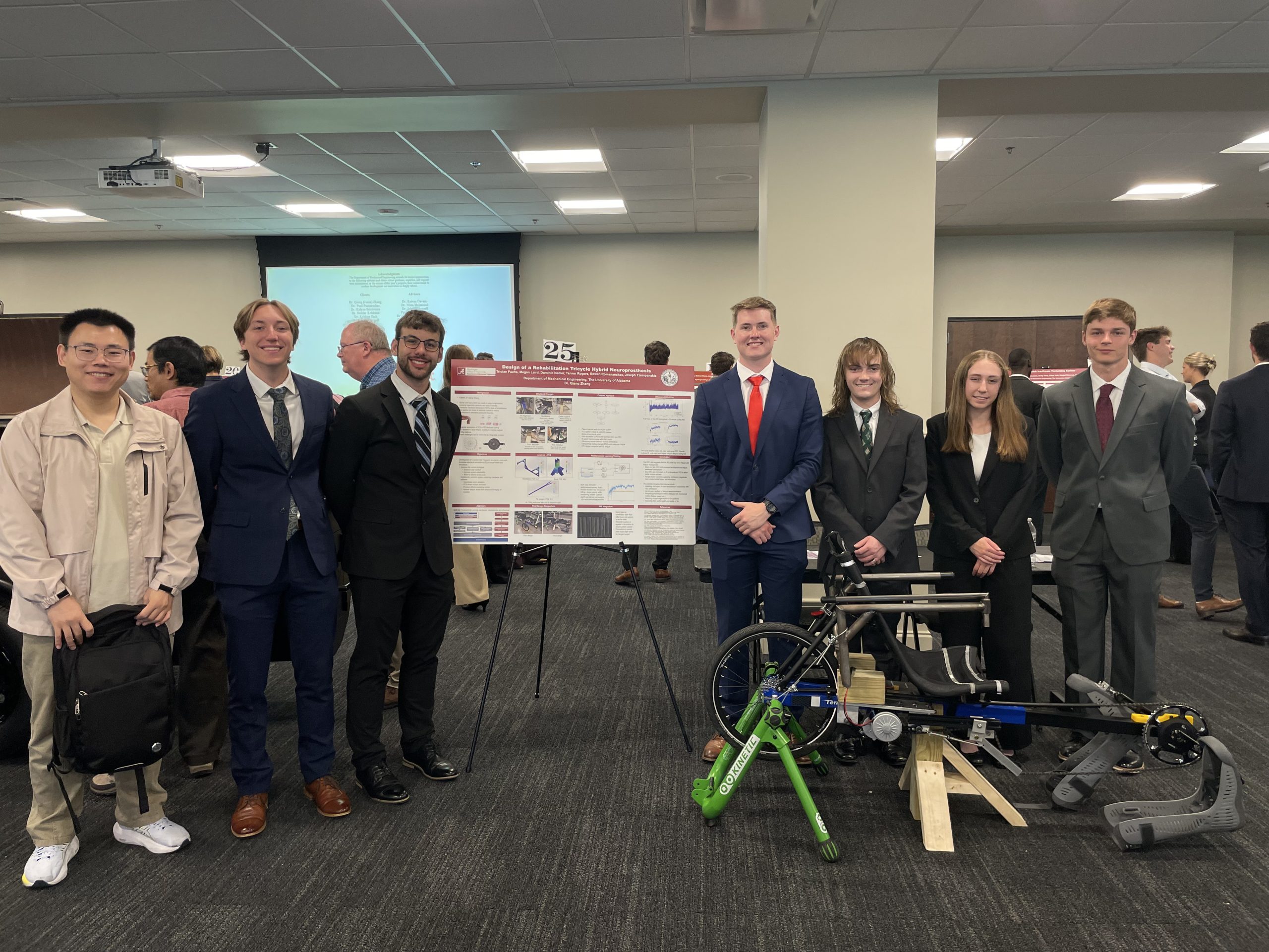 Six people stand in business attire next to a project display and a mechanical device, possibly a bike-related engineering project, in a conference room with other groups and posters visible in the background.