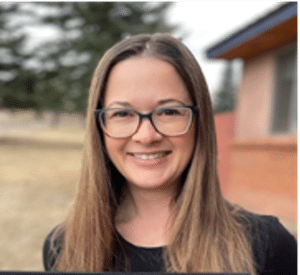 A woman with long straight brown hair and glasses smiles outdoors. She is wearing a black top, with trees and a building visible in the blurred background.