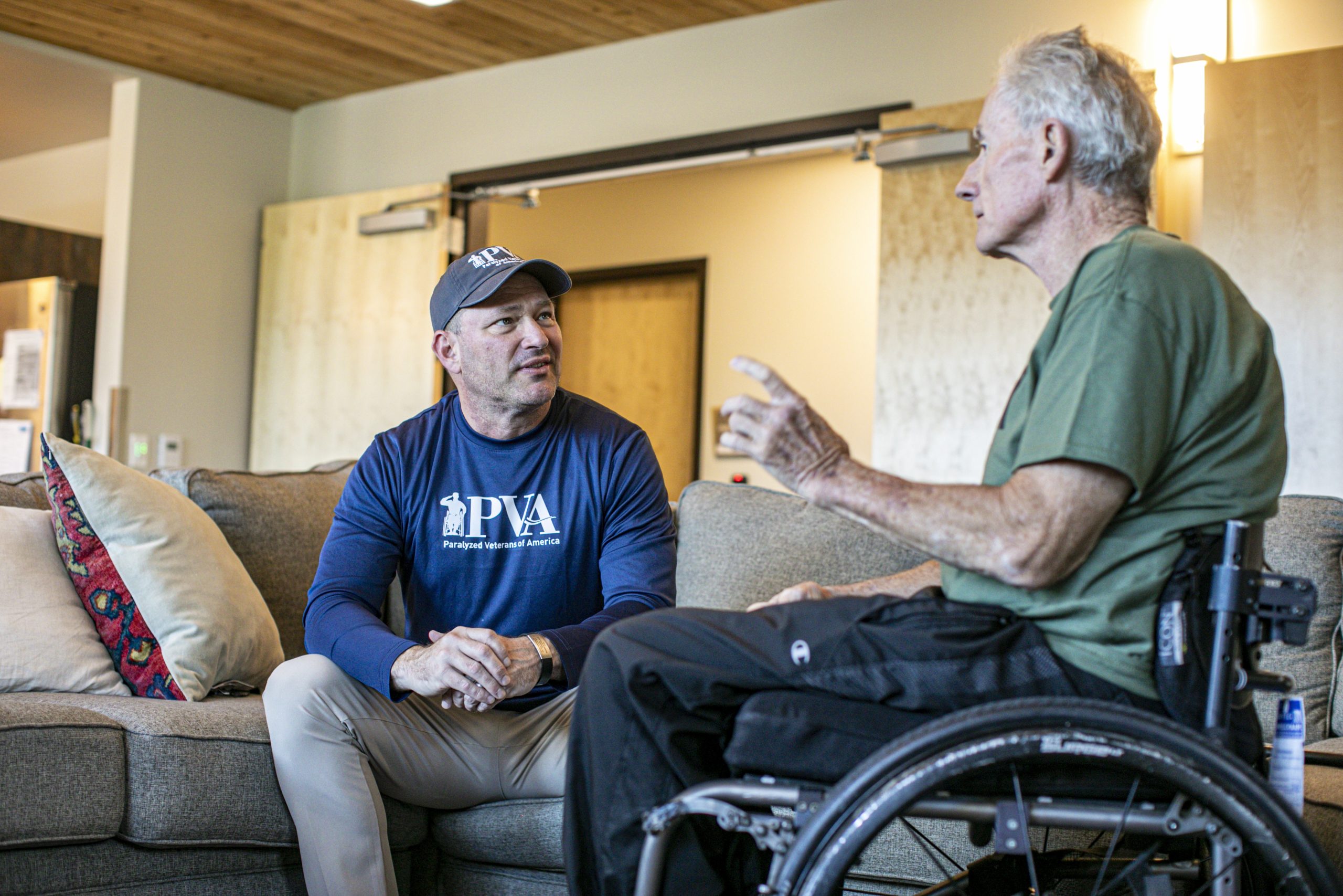 Two men talk in a living room. One man in a blue PVA shirt and baseball cap sits on a couch, while the other, wearing a green shirt, sits in a wheelchair and gestures with his hand.