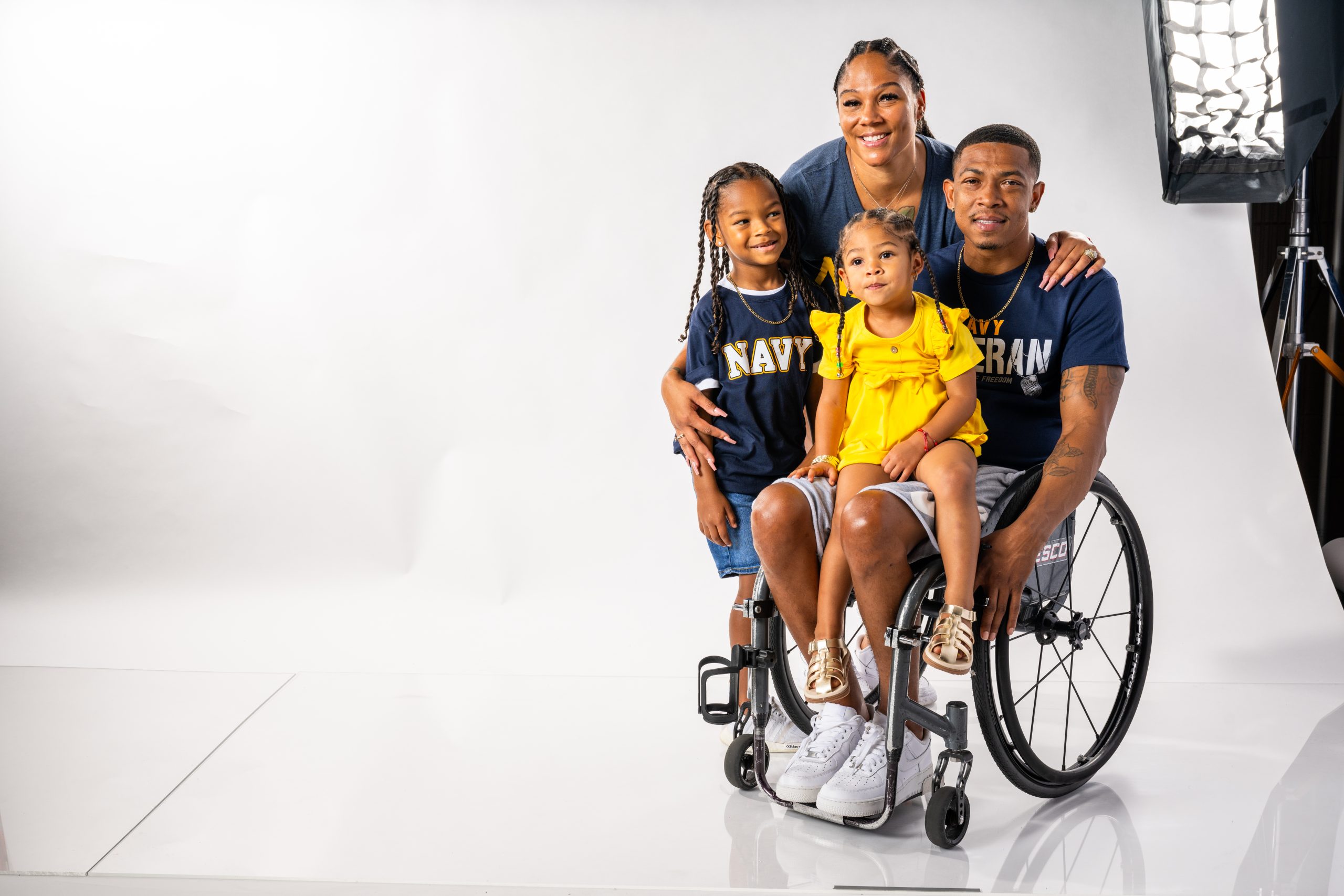 A smiling family of four poses in a photo studio. One adult male in a wheelchair and young girl sit in front. An adult female and another young girl stand behind them. The adults wear matching "NAVY" and "VETERAN" shirts.