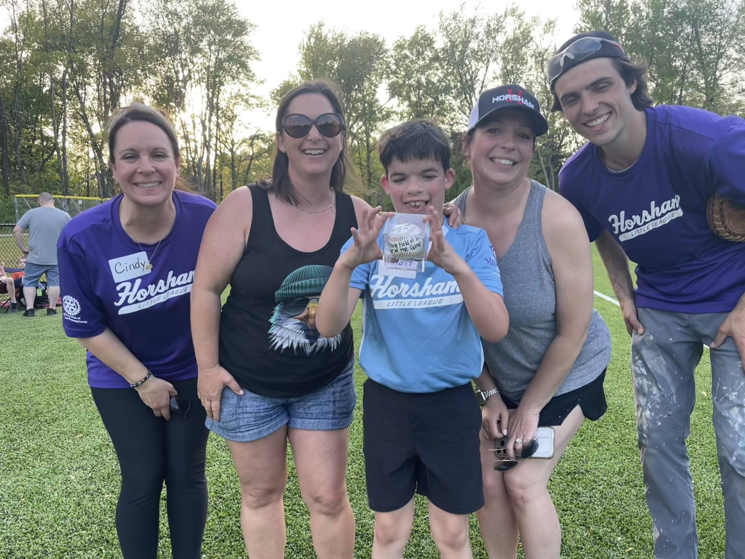 A smiling boy holds a baseball as he stands with four adults on a grassy field. Everyone is smiling at the camera, and some are wearing shirts that say "Horsham." Trees and sunlight are visible in the background.