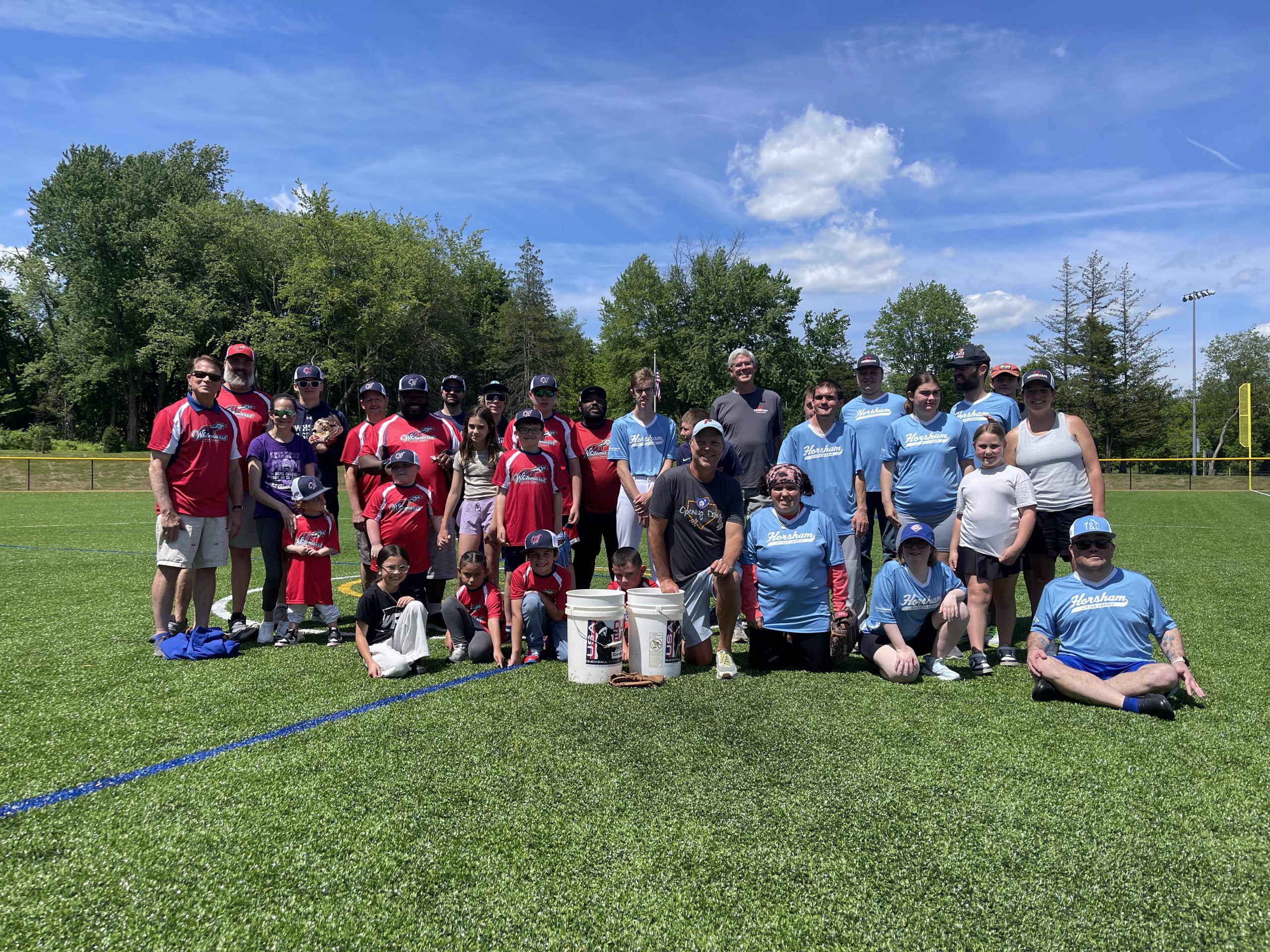 A group of kids and adults pose together on a sunny baseball field, wearing red and blue team shirts. Some kneel or sit in front, while others stand behind, with trees and a blue sky in the background.