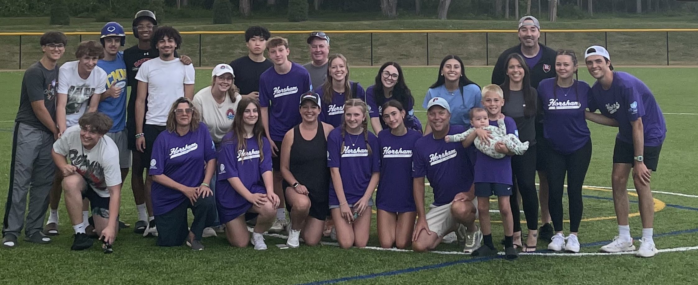 A group of people, many wearing matching purple shirts, pose and smile together on a sports field with trees and a yellow fence in the background. Some kneel in front, while others stand behind them.