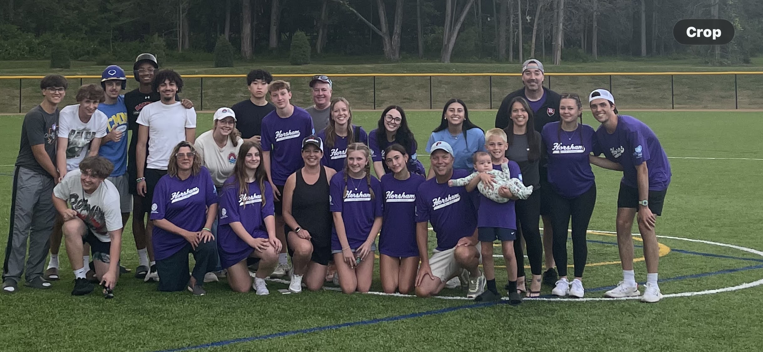 A group of people, many wearing matching purple shirts, pose and smile together on a sports field with trees and a yellow fence in the background. Some kneel in front, while others stand behind them.