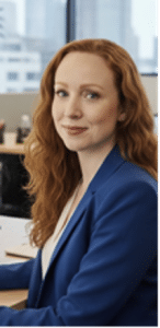 A woman with long red hair, wearing a blue blazer, sits at a desk in a modern office, looking at the camera with a gentle smile.