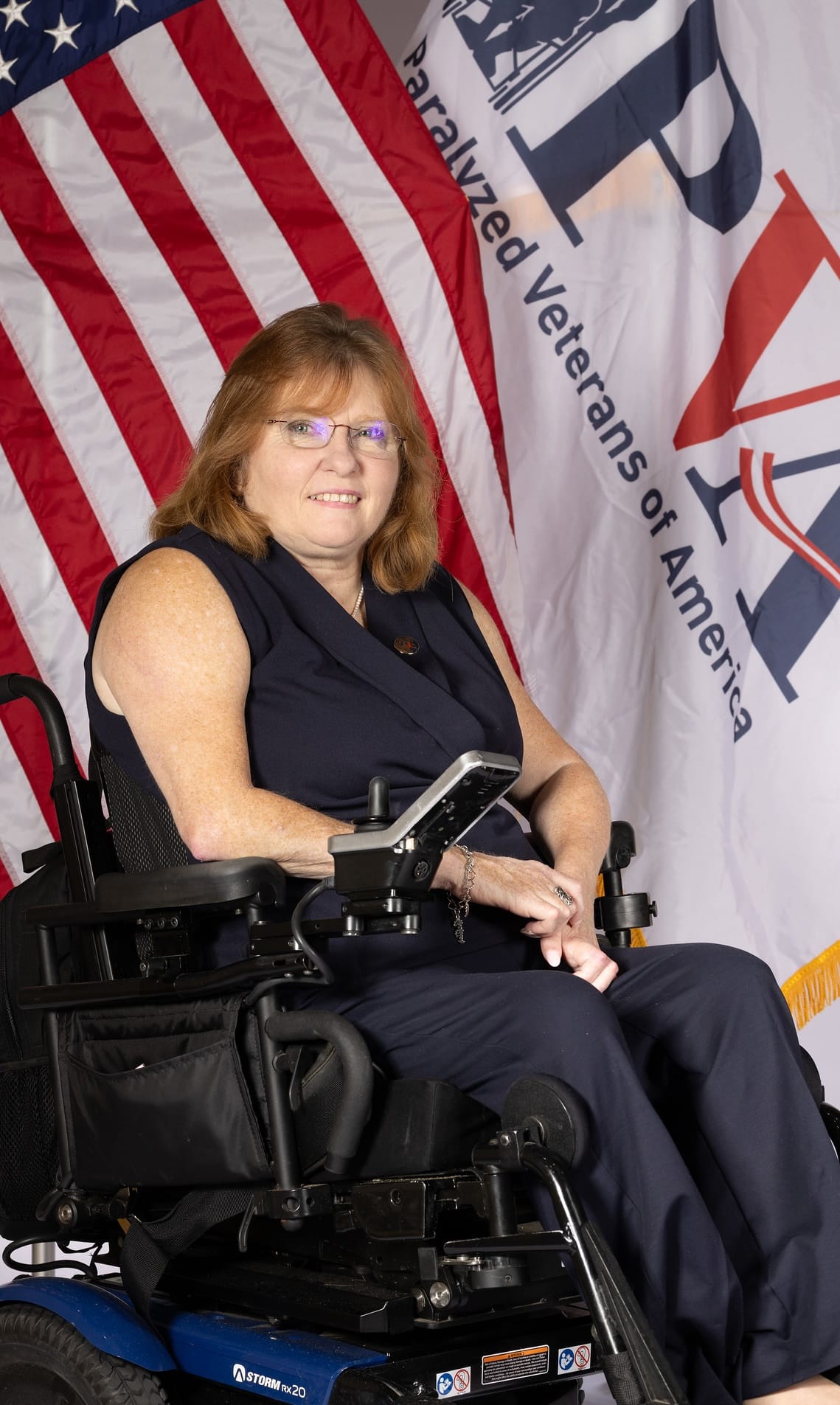 A woman with long, light brown hair and glasses is sitting in a wheelchair. She is wearing a maroon outfit and smiling. Behind her is an American flag and a large banner with the text "Paralyzed Veterans of America".