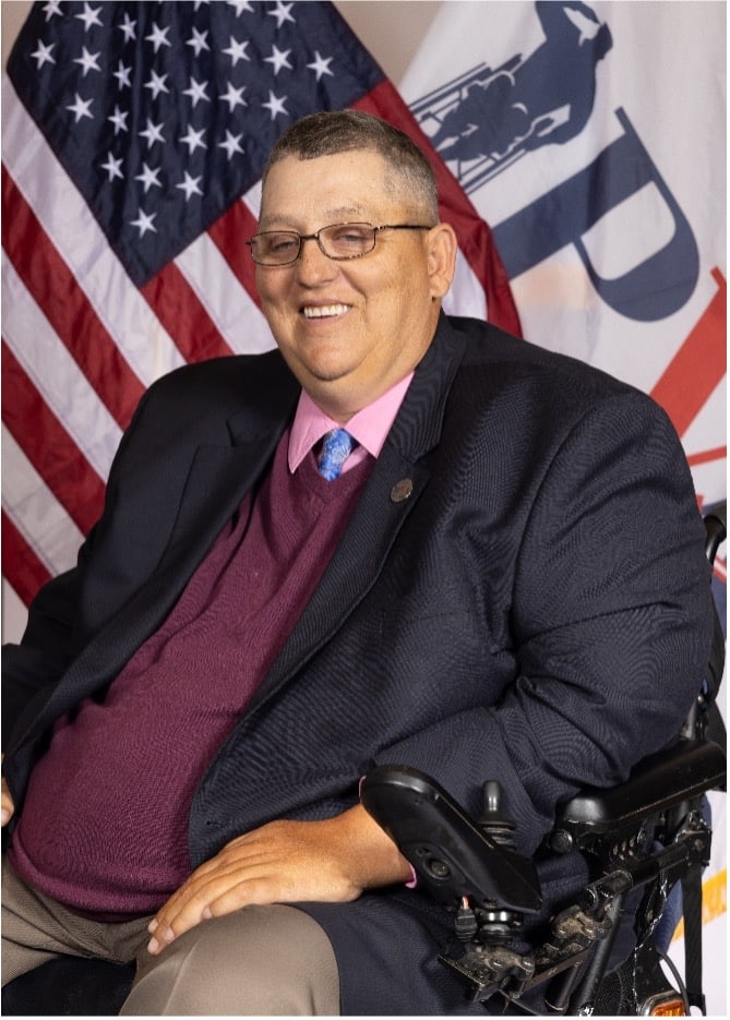 A man in a suit, pink shirt, and maroon sweater sits in a wheelchair, smiling. Behind him are a U.S. flag and a white flag with partially visible lettering and a graphic.