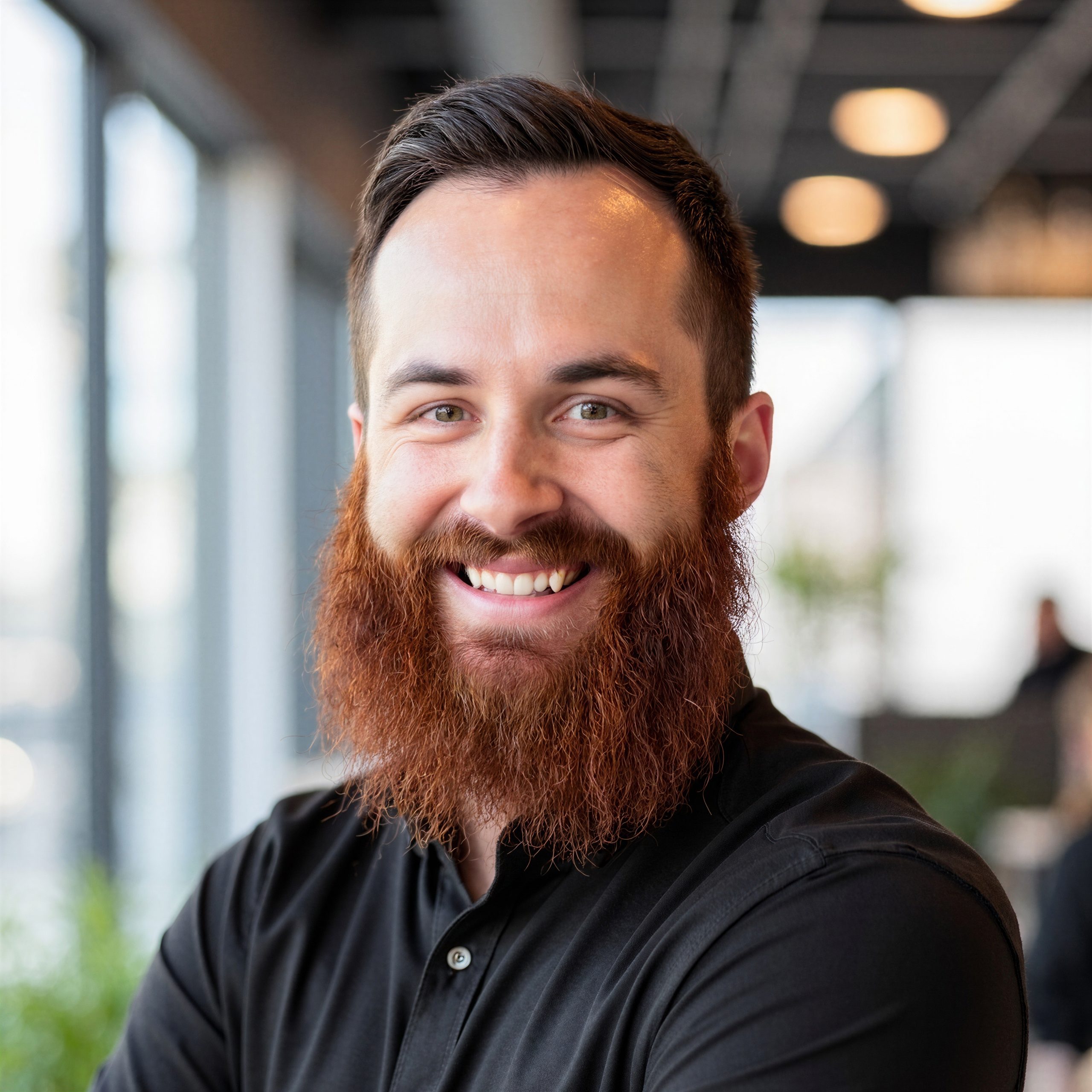 A man with a long red beard and short brown hair smiles while standing indoors. He is wearing a black button-up shirt and is in a modern, bright office setting with blurred lights and windows in the background.