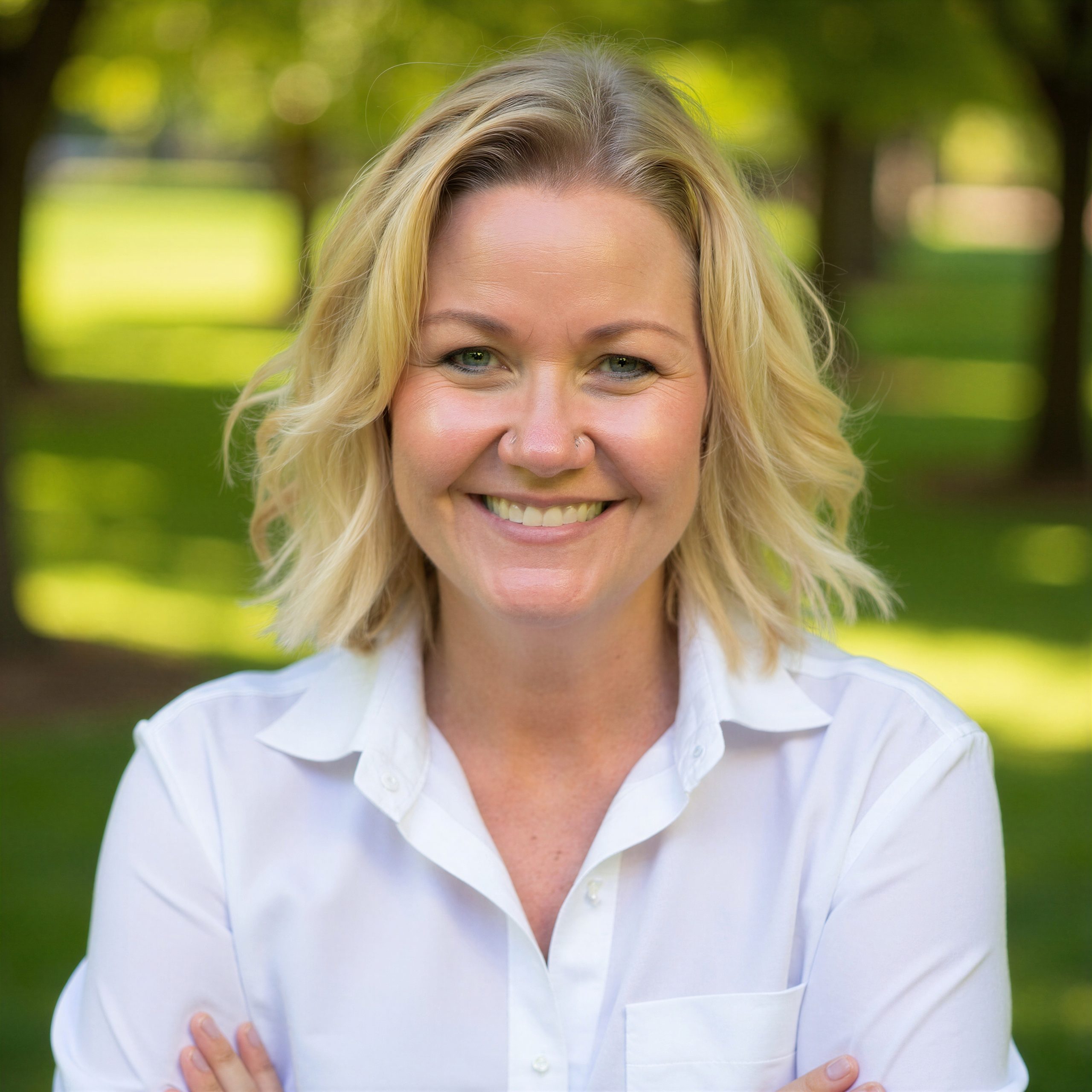 A smiling woman with blonde hair wearing a white shirt stands outdoors with green trees and grass in the background. She has her arms crossed and looks directly at the camera.