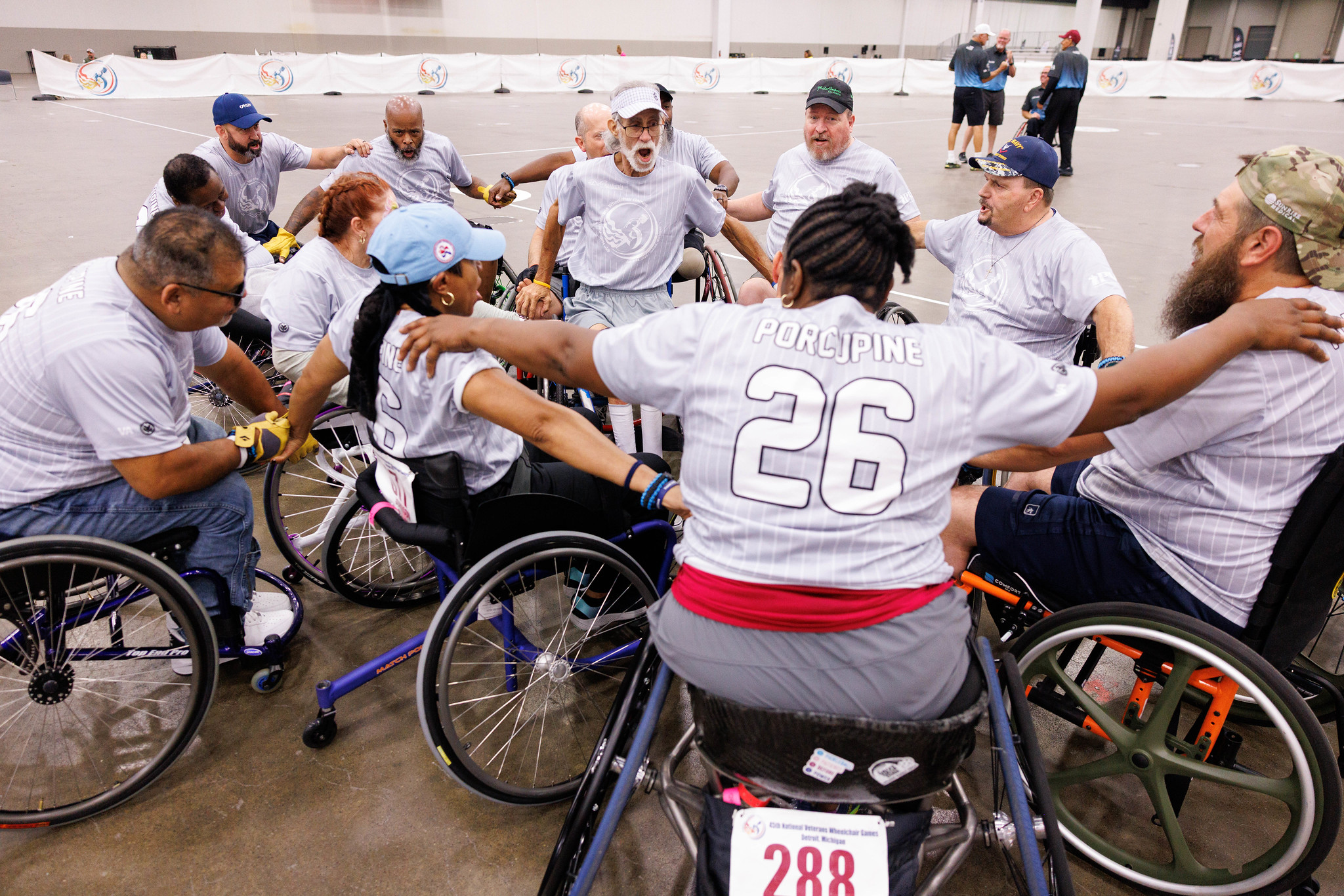 Two men in wheelchairs smile at the camera in an indoor event space. One wears a white T-shirt with a PVA logo; the other wears a navy shirt and backpack. Others and tables with laptops are visible in the background.