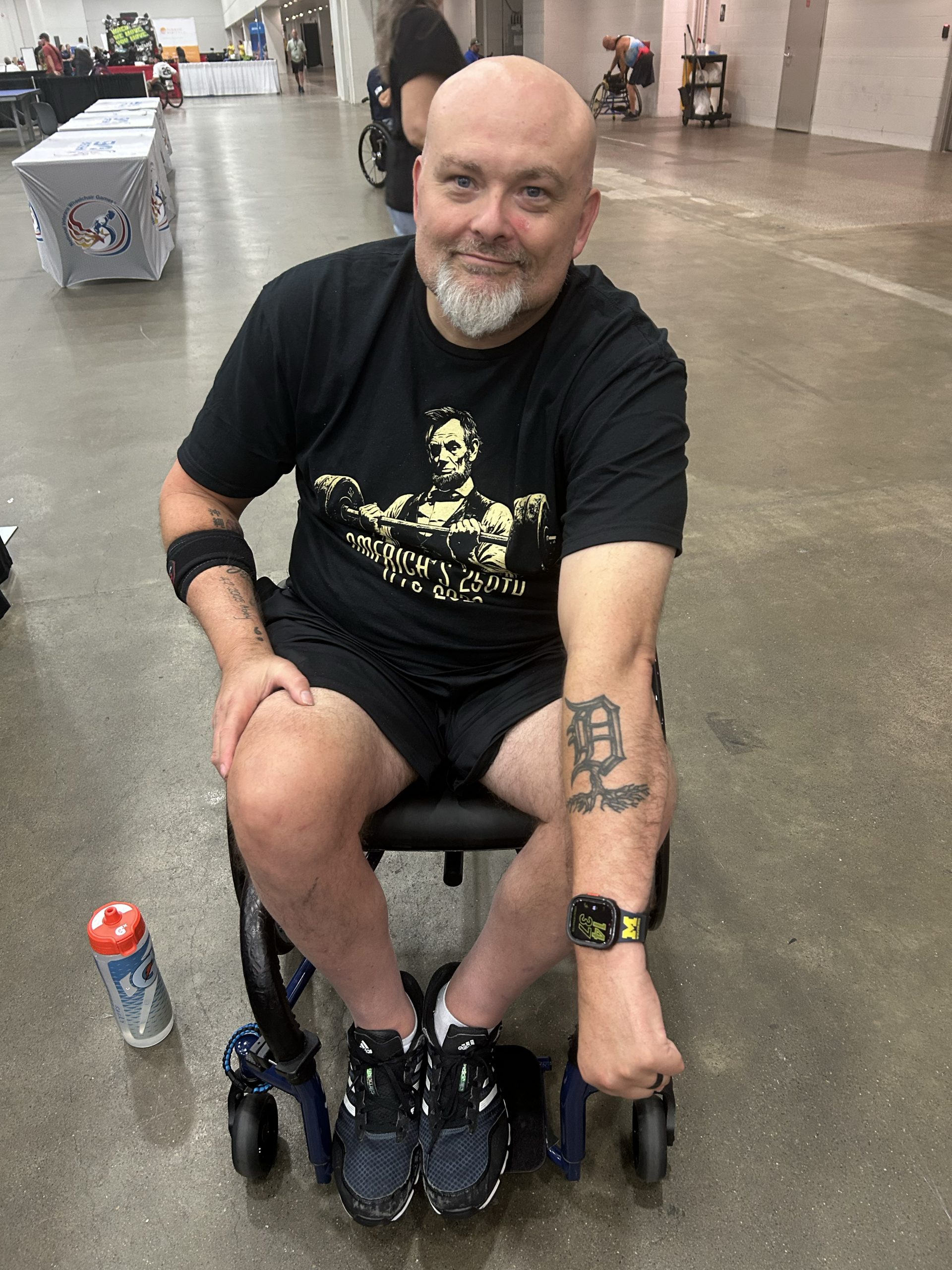 A man with a beard and tattoos sits in a wheelchair. He is wearing a black T-shirt, shorts, and sneakers. The background shows a large indoor space with a few people and objects visible.