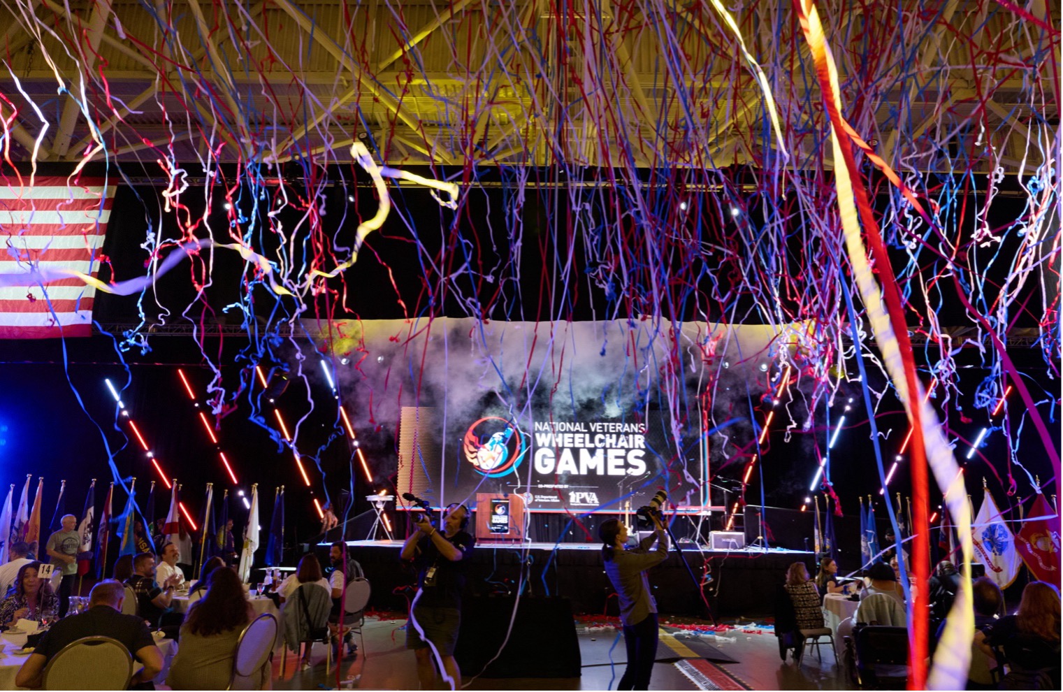 Colorful streamers fall over a stage with "National Veterans Wheelchair Games" displayed on a screen. People sit at tables and a large American flag hangs on the left side of the image.