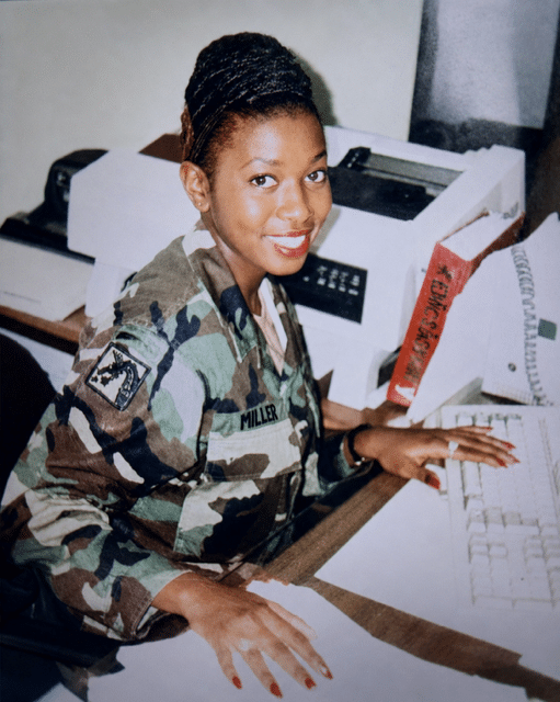A woman in military uniform sits at a desk, smiling at the camera, with her hands on a computer keyboard. Behind her are a printer and papers.