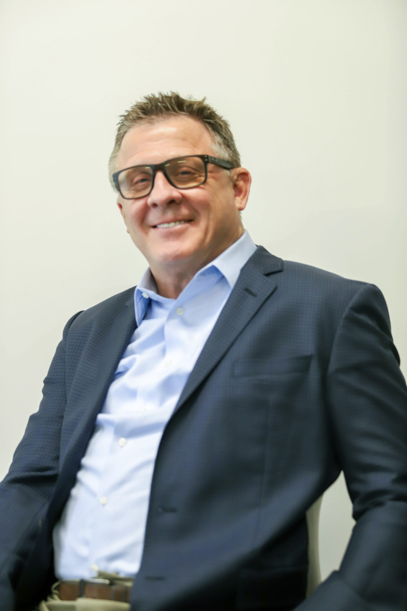 A man in a dark suit jacket and light blue shirt sits and smiles at the camera; he wears glasses and is posed against a plain light-colored background.