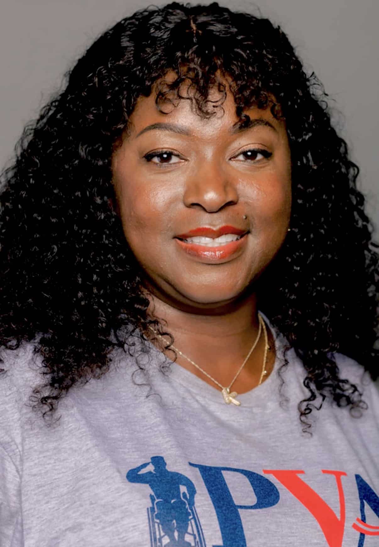 A woman with curly black hair smiles at the camera, wearing a gray T-shirt with colorful letters and a gold necklace on a plain background.
