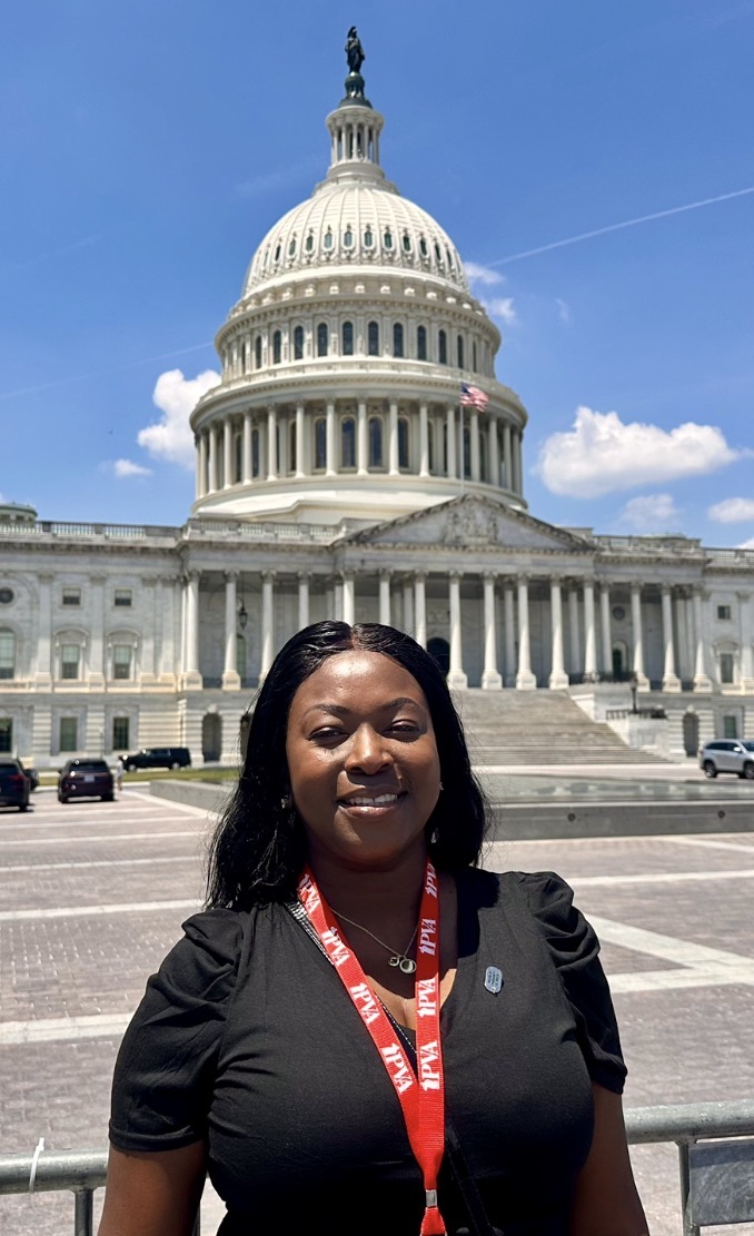 A woman wearing a black shirt and red lanyard stands smiling in front of the U.S. Capitol building on a sunny day with a blue sky and scattered clouds.