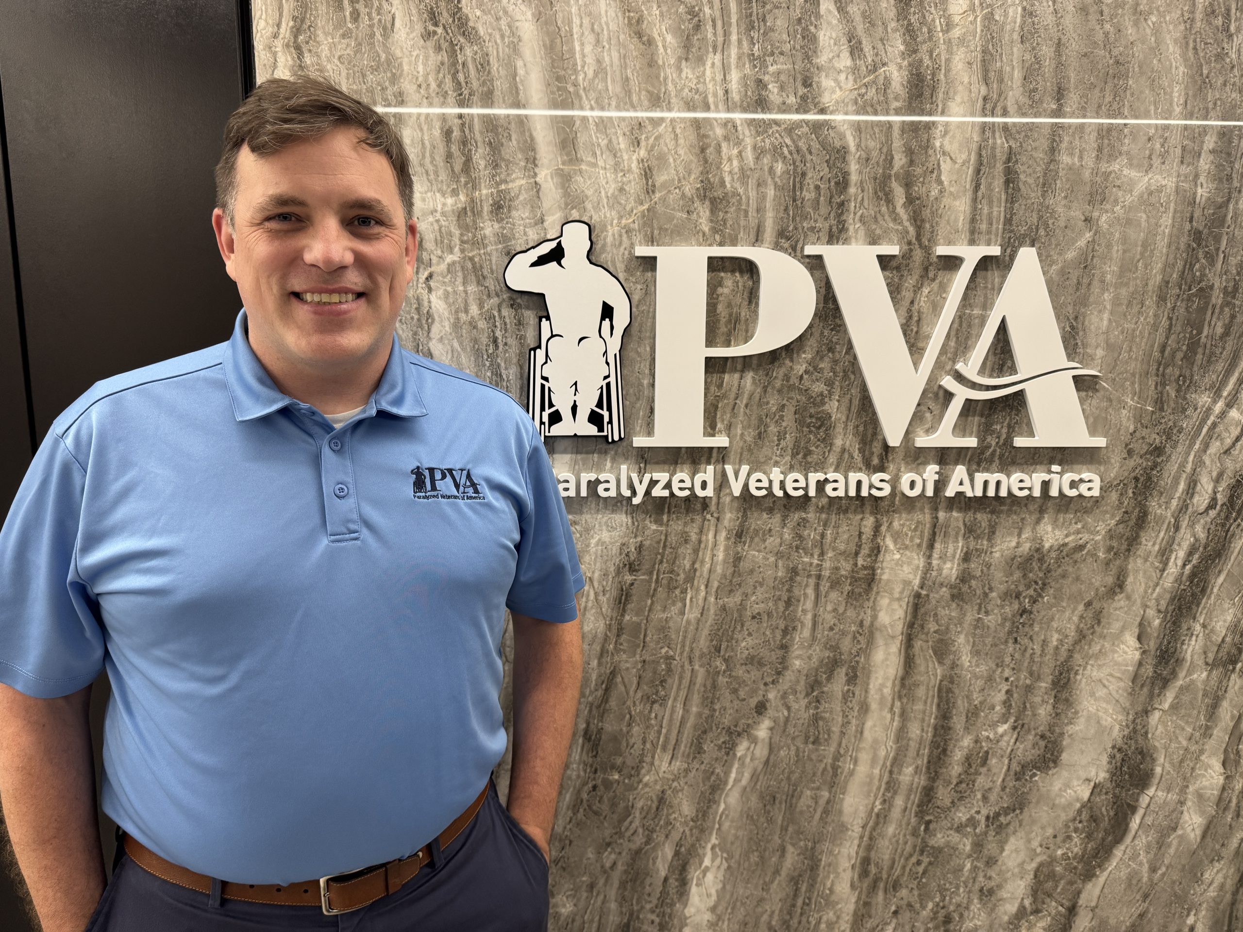 A man in a light blue polo shirt stands smiling next to a wall with the Paralyzed Veterans of America (PVA) logo and text on it.