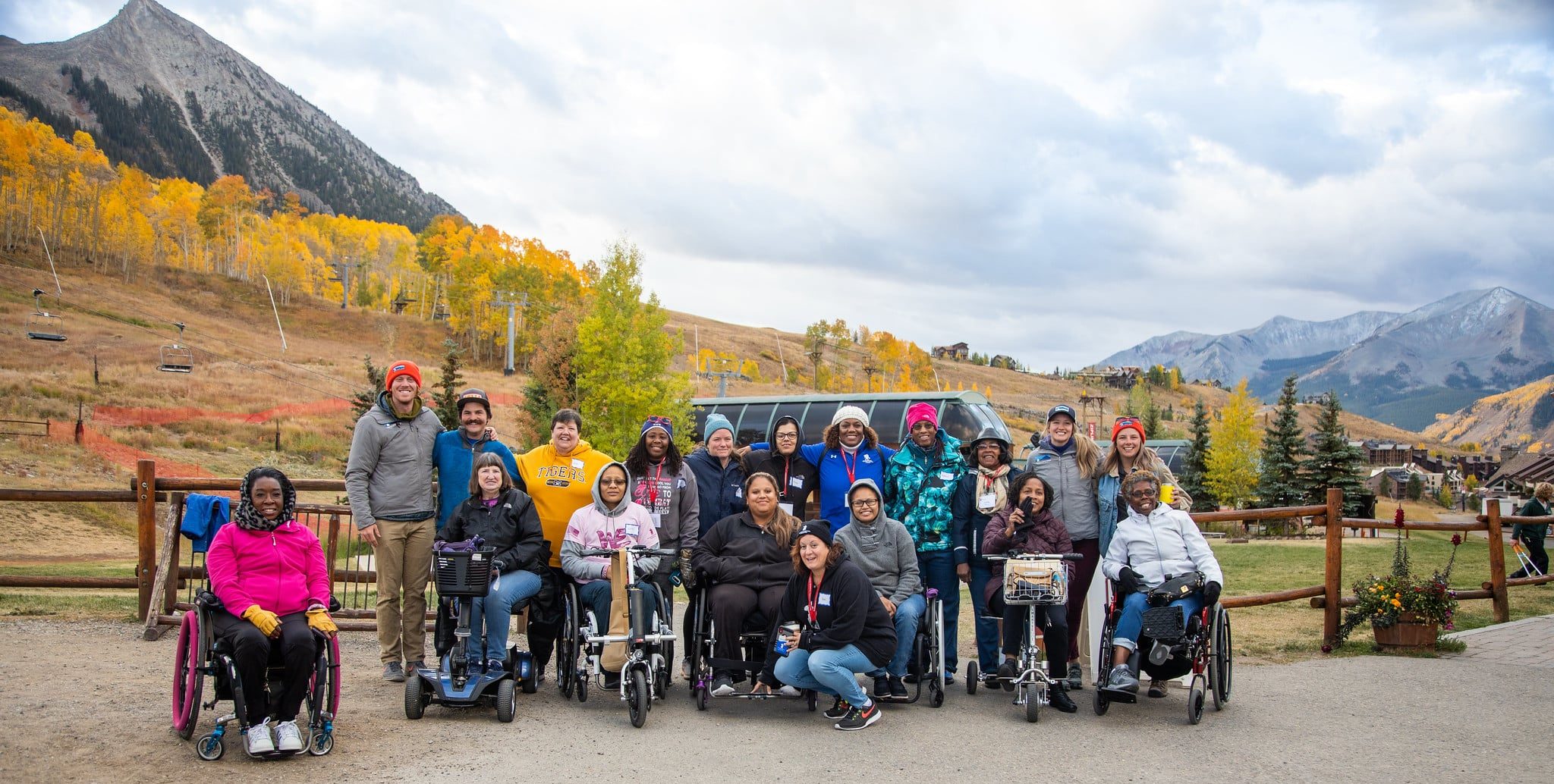 A diverse group of people, some in wheelchairs, pose and smile outdoors on a paved road with autumn trees, mountains, and a wooden fence in the background.