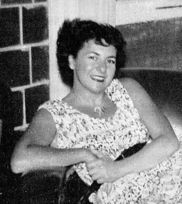 A woman with dark, curly hair in a light-patterned dress sits smiling on a chair near a fireplace. The photo is black and white, and has a vintage appearance.